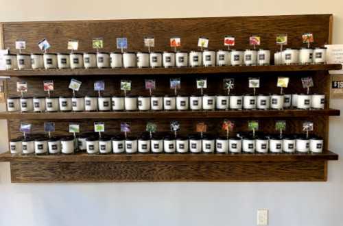 A display of white candles on wooden shelves, each labeled with colorful scent tags.