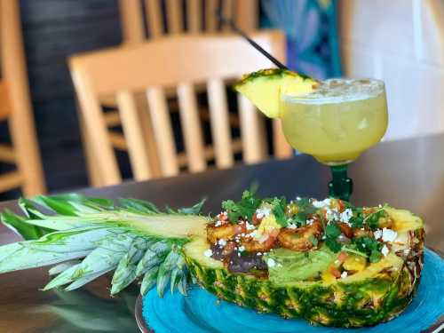 A vibrant pineapple dish filled with shrimp, avocado, and toppings, alongside a refreshing cocktail.