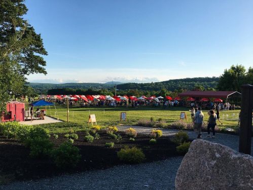A vibrant outdoor event with red tents, people mingling, and a scenic view of mountains in the background.