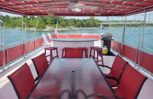 A spacious boat deck with a large table, red chairs, a bar area, and a view of the water and trees.