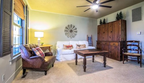 Cozy living room with a white sofa, wooden table, leather chair, and decorative accents. Warm lighting and soft carpet.
