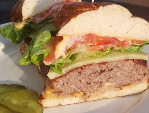 A close-up of a sliced burger on a pretzel bun, layered with lettuce, tomato, cheese, and a side of pickles.