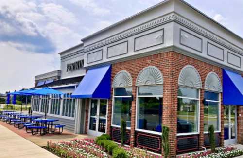 Exterior of a restaurant with blue awnings, large windows, and a landscaped area with flowers and seating.