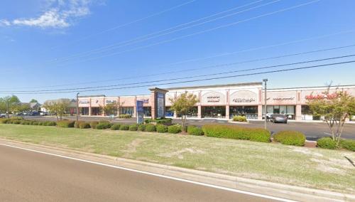 A row of retail stores along a road, with green grass and trees in the foreground under a clear blue sky.