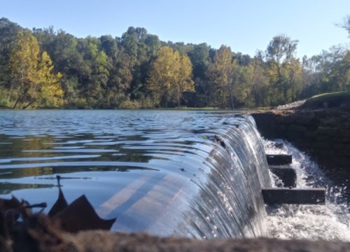 A serene river scene with a small waterfall, surrounded by trees and clear blue skies.