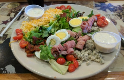 A colorful salad with greens, cherry tomatoes, hard-boiled eggs, ham, bacon, cheese, and two dressings on the side.