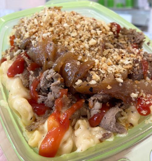 A green container filled with macaroni and cheese topped with shredded beef, caramelized onions, ketchup, and breadcrumbs.
