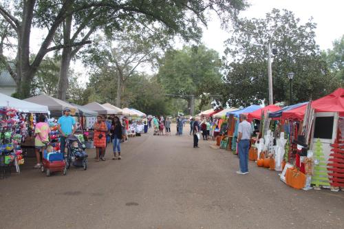 A bustling outdoor market with colorful tents, vendors, and visitors along a tree-lined street.