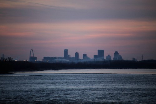 Silhouette of a city skyline at dusk, with a river in the foreground and a colorful sky.