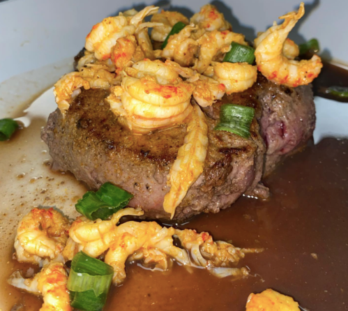 A close-up of a cooked steak topped with shrimp and green onions, served in a rich brown sauce.