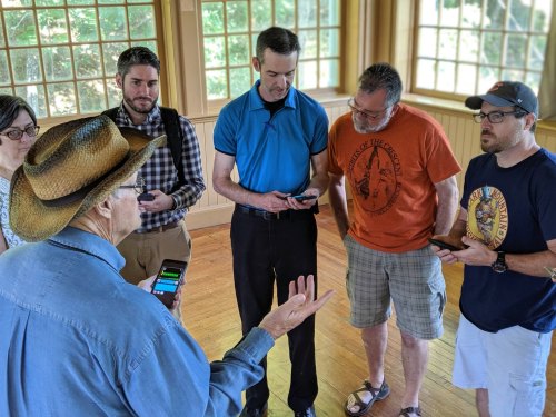 A group of people in a room, gathered around a man in a hat, engaged in discussion while using their phones.