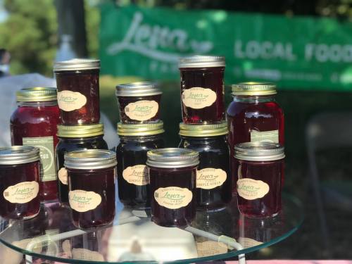 A display of jars filled with various jams and preserves, labeled and arranged on a table with a green "Local Food" banner in the background.
