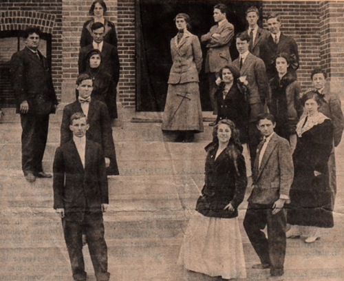 A historical group photo of men and women in early 20th-century attire, standing outside a brick building.