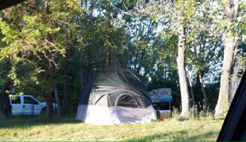 A large tent set up in a grassy area surrounded by trees, with a white vehicle parked nearby.