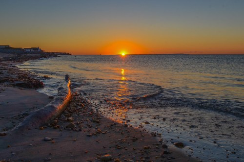 Sunset over a calm beach, with gentle waves lapping at the shore and a log resting on the sand.