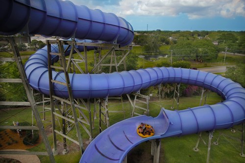 Aerial view of a purple water slide twisting down from a tall structure, surrounded by greenery and a cloudy sky.