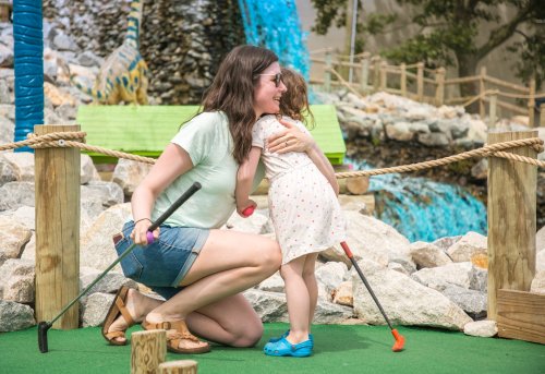 A woman hugs a young girl on a mini-golf course, surrounded by rocks and a waterfall backdrop.