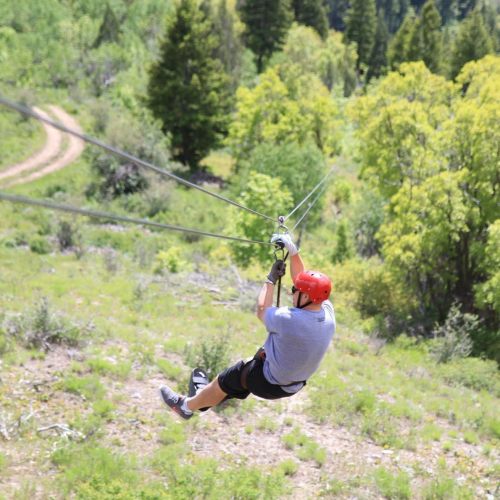 A person in a red helmet zip-lining over a green landscape with trees and a dirt path below.