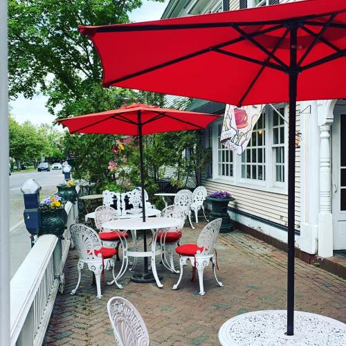 Outdoor dining area with white furniture and red umbrellas, surrounded by greenery and flowers.