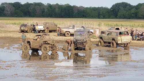 Off-road vehicles stuck in mud, with people assisting and onlookers in the background, surrounded by a rural landscape.