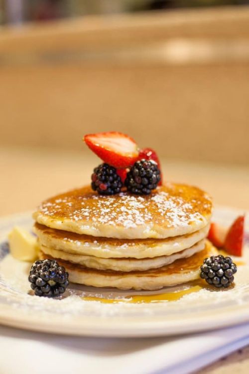 A stack of fluffy pancakes topped with strawberries and blackberries, dusted with powdered sugar and served with butter.