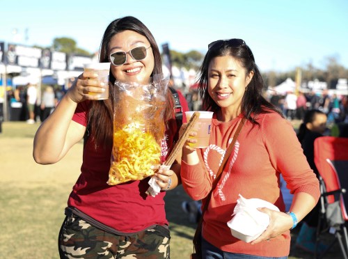 Two women smile and pose with snacks and drinks at an outdoor event, surrounded by festival tents in the background.