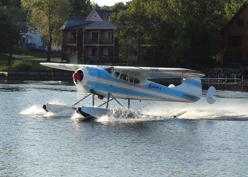 A seaplane with blue stripes takes off from a calm lake, creating splashes of water around it.