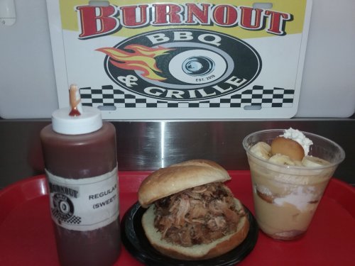 A BBQ sandwich with pulled pork, a bottle of sauce, and a dessert cup with banana pudding on a red tray.