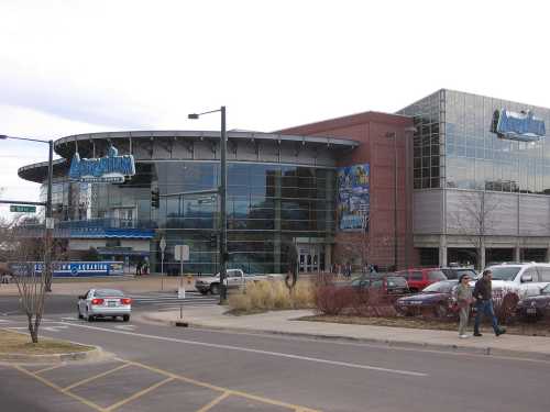 Exterior view of a modern sports arena with large glass windows and a brick facade, featuring people walking outside.