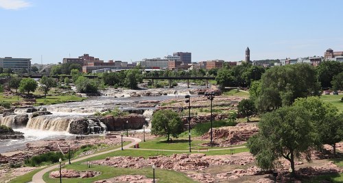 A scenic view of waterfalls and a park with city buildings in the background under a clear blue sky.
