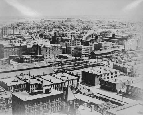 Aerial view of a historic cityscape featuring buildings, rooftops, and railway tracks in black and white.