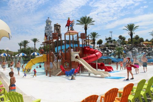 A vibrant water park with a large play structure, slides, and families enjoying the sunny day. Palm trees in the background.