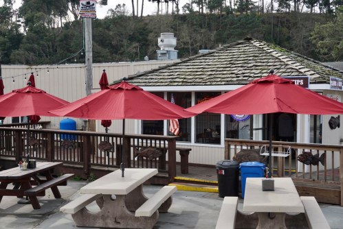 Outdoor dining area with picnic tables and red umbrellas in front of a casual restaurant with a wooden deck.