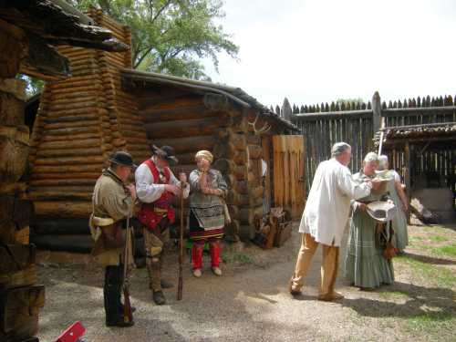 A group of people in historical costumes gather outside log cabins, engaging in conversation and activities.