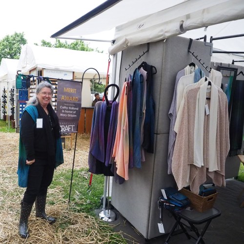 A smiling woman stands in front of a display of colorful textiles at an outdoor craft fair.