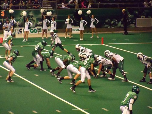 A football game in progress, with players in green and white uniforms on a green field, cheerleaders in the background.