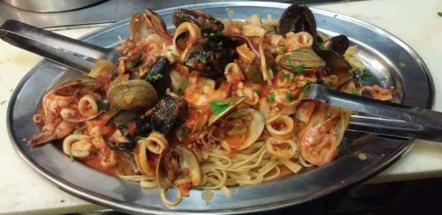 A large platter of seafood pasta with clams, shrimp, and squid in a rich tomato sauce, garnished with herbs.