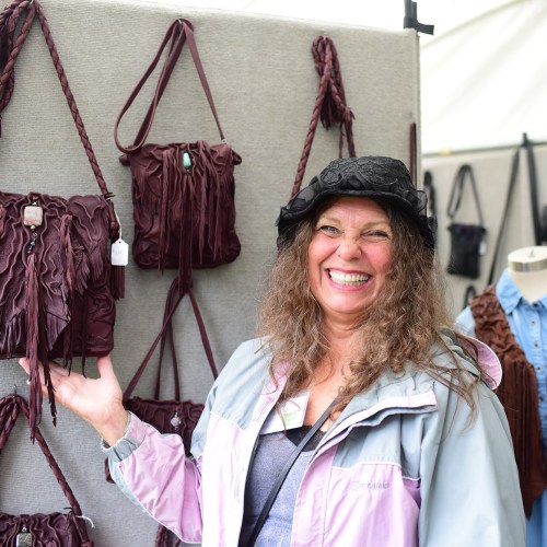 A smiling woman in a hat gestures towards leather bags displayed on a wall at a market.