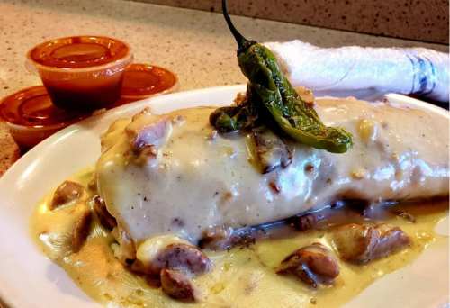 A large burrito smothered in creamy sauce, topped with a green chili, served with two small cups of salsa.