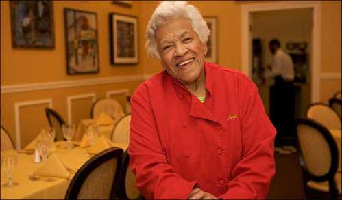 A smiling elderly woman in a red chef's coat stands in a warmly decorated restaurant.