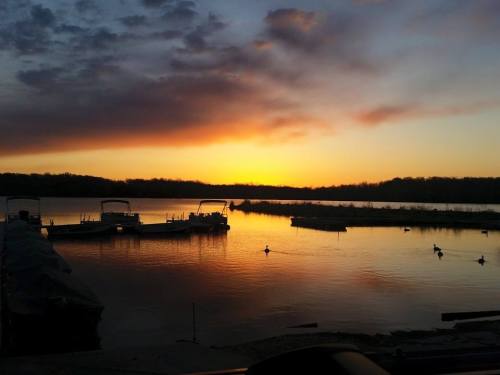Sunset over a calm lake with boats docked and ducks swimming, casting a warm glow on the water.