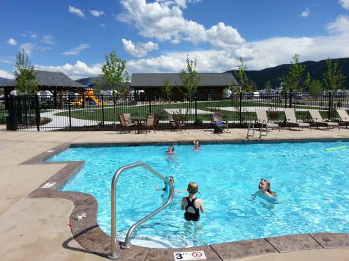 A sunny pool scene with children swimming, surrounded by lounge chairs and trees, with playground equipment in the background.