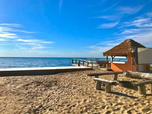 A sandy beach with a thatched-roof hut and a wooden pier extending into a calm blue sea under a clear sky.