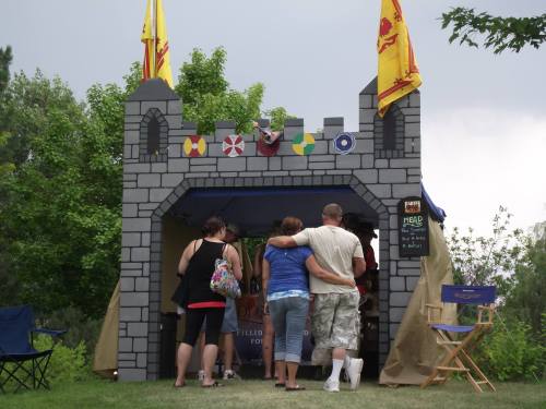 A group of people approaches a castle-themed booth with flags and banners in a park setting.