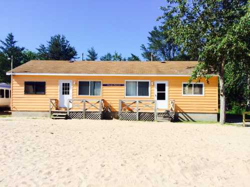 A bright orange beach house with two entrances, surrounded by sand and trees under a clear blue sky.