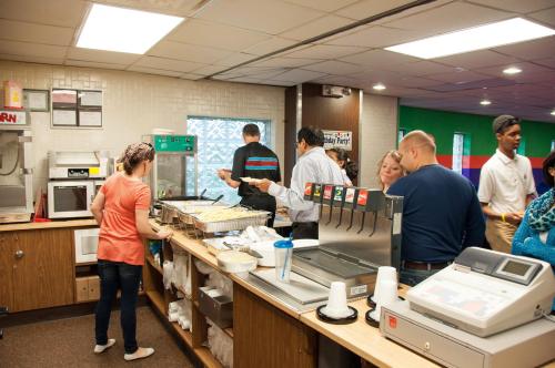 A busy cafeteria with people serving food, chatting, and using a cash register in a bright, casual setting.