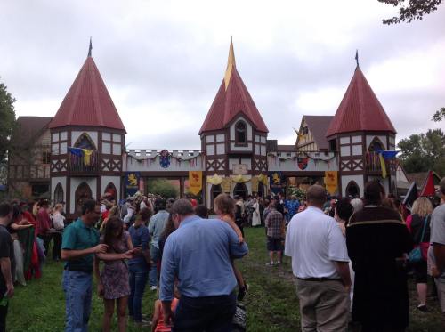 A crowd gathers in front of a medieval-style building with red roofs and banners, set against a cloudy sky.