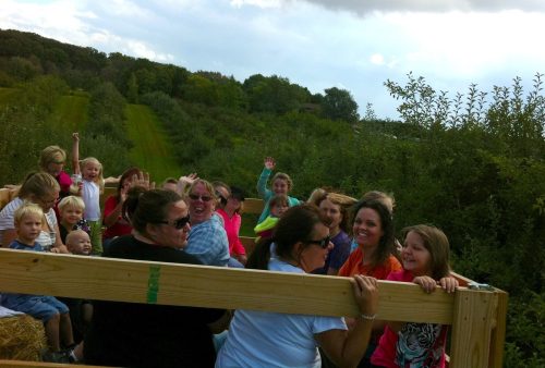 A group of children and adults smiling and waving while riding on a wooden wagon through an orchard.