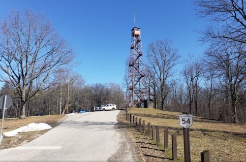 A gravel road leads to a tall observation tower surrounded by bare trees under a clear blue sky. A sign reads "54."