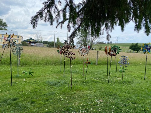 Colorful metal flowers on tall stakes in a grassy field, with trees and a distant building under a cloudy sky.